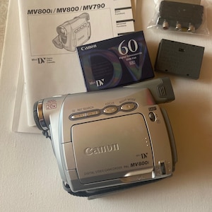 May include: A silver Canon MV800i digital video camcorder with a black strap. The camcorder has a 20x zoom lens, a record search button, a focus button, a night mode button, and an easy button. The camcorder is on a white surface.