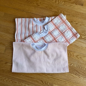 Puede incluir: Tres baberos para bebés en tonos rosa y blanco. Un babero tiene rayas verticales, otro un estampado de cuadros y el tercero rayas horizontales. Cada babero tiene un cuello blanco.