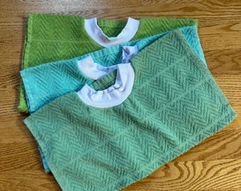 May include: Three terry cloth bibs in shades of green and blue, each with a white collar. The bibs are arranged on a wooden surface, showcasing their textured, patterned design. The bibs are a practical and stylish accessory for mealtime.