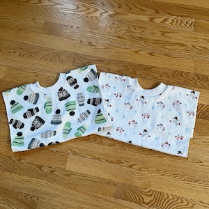 May include: Two white bibs with different winter-themed patterns. One bib has a pattern of green and black mittens and hats. The other bib has a pattern of snowmen and snowflakes.