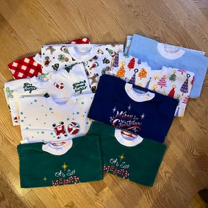 May include: A collection of ten Christmas-themed bibs in various colors and patterns. The bibs feature festive designs such as candy canes, Christmas trees, Santa Claus, and gingerbread men. The text on the bibs includes "Gimme some sugar", "Merry Christmas", and "Oh So Merry".