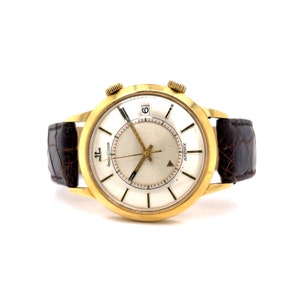 May include: A gold-tone wristwatch with a white face and black hands. The watch has a brown leather band and the words "Automatic" and "Jaeger-LeCoultre" are printed on the face.