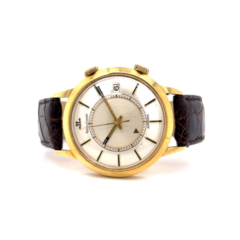 May include: A gold-tone wristwatch with a white face and black hands. The watch has a brown leather band and the words "Automatic" and "Jaeger-LeCoultre" are printed on the face.