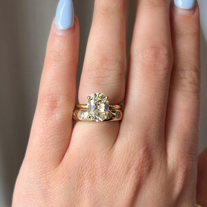Pode incluir: Um close-up de uma m&atilde;o usando uma banda de ouro com um grande anel de diamante redondo e claro. O anel &eacute; engastado em uma banda de ouro com uma borda delicada de milgrain. A m&atilde;o tamb&eacute;m est&aacute; usando uma banda de ouro fina com pequenos diamantes engastados em pav&eacute;.