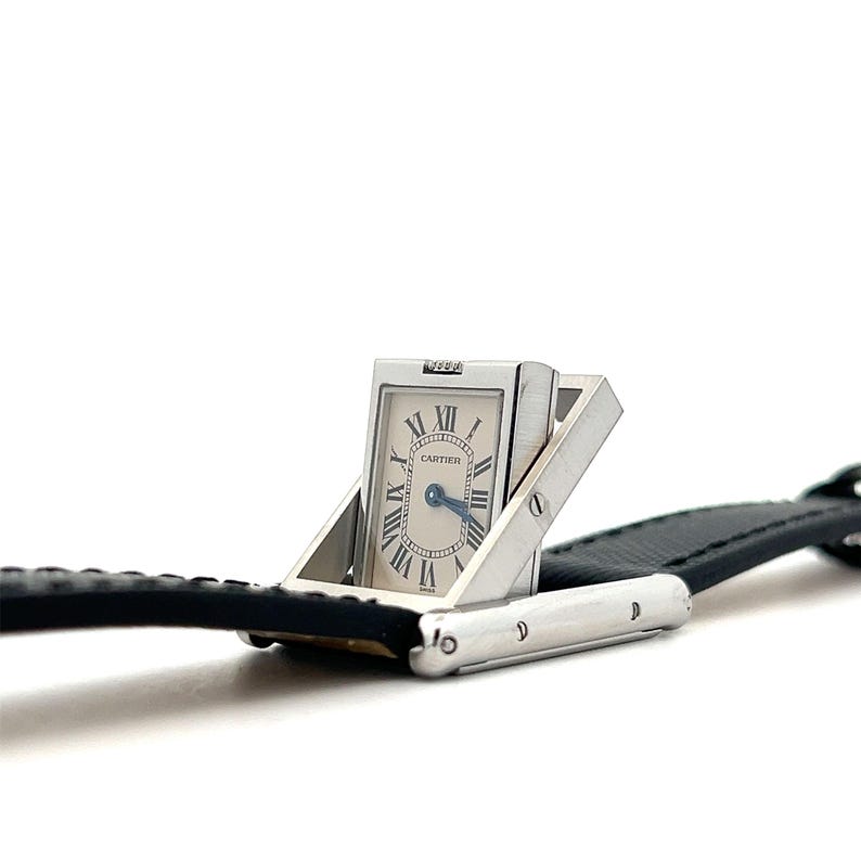 May include: Close-up view of a Cartier wristwatch with a unique folding design. The watch face is rectangular with Roman numerals and a Cartier logo. The watch has a silver-toned metal body and a black leather strap. The watch's unique feature is its ability to fold, revealing the watch face when opened.