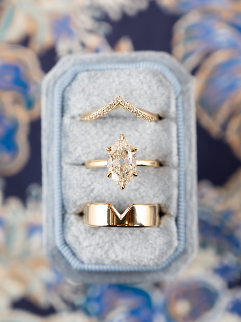 May include: Three gold rings with diamonds in a blue velvet ring box. The top ring is a delicate band with a chevron shape of diamonds. The middle ring features a large, pear-shaped diamond. The bottom ring is a thick band with a V-shaped cutout.