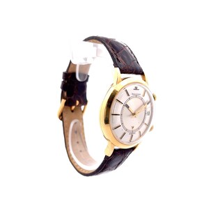 May include: A gold-tone wristwatch with a white face and black leather band. The watch has a date window and a second hand. The watch is made by Jaeger-LeCoultre.