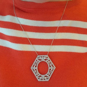 May include: A silver necklace with a hexagonal pendant featuring a detailed, ornate design with a circle in the center. The necklace is worn over a red and white striped sweater.