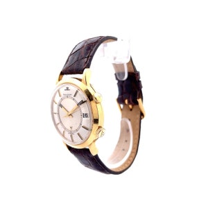 May include: A gold-tone wristwatch with a white face and black hands. The watch has a brown leather band and a date window at the 3 o'clock position.