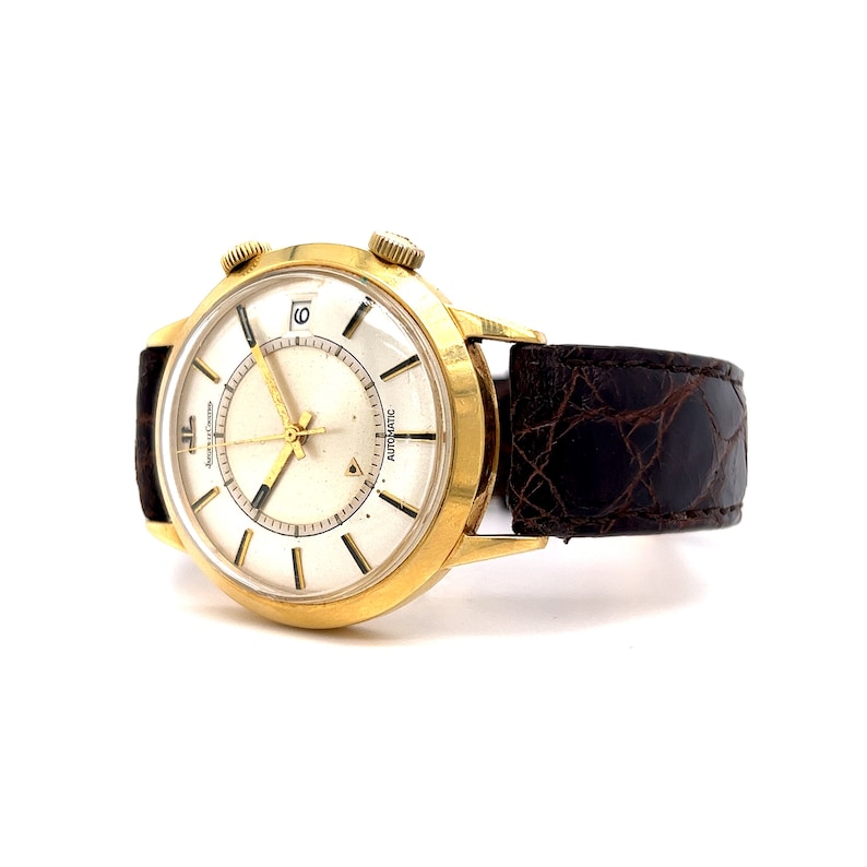 May include: A gold-tone wristwatch with a white face and black leather band. The watch has a date window at the 3 o'clock position and the words "Automatic" and "Jaeger-LeCoultre" printed on the face.