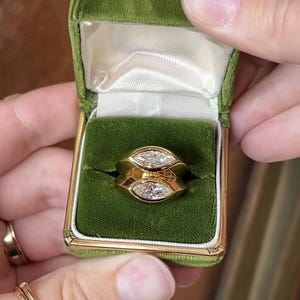 Pu&ograve; includere: Un anello d'oro con due diamanti a forma di marquise incastonati in una montatura a bezel. L'anello &egrave; in una scatola per anelli in velluto verde.