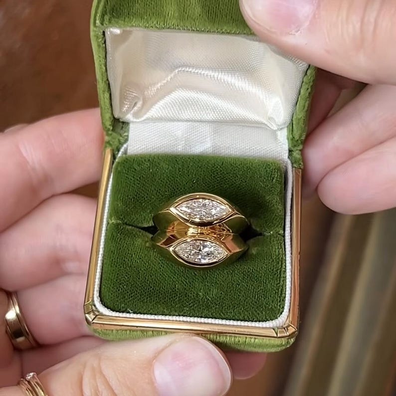 Pu&ograve; includere: Un anello d'oro con due diamanti a forma di marquise incastonati in una montatura a bezel. L'anello &egrave; in una scatola per anelli in velluto verde.