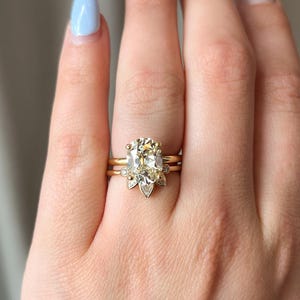 Pode incluir: Um anel de ouro com uma grande pedra de diamante central de forma oval, cercada por diamantes menores. O anel &eacute; usado em um dedo com esmalte de unha azul claro.