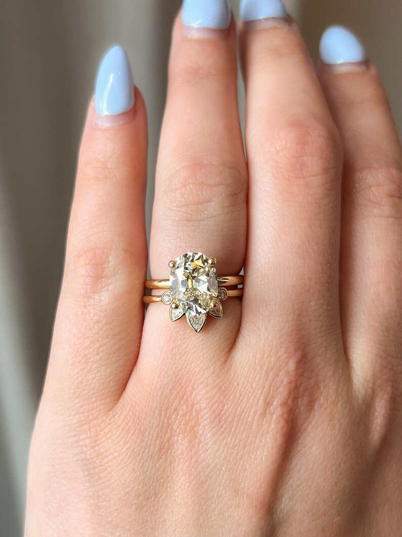Pode incluir: Um anel de ouro com uma grande pedra de diamante central de forma oval, cercada por diamantes menores. O anel &eacute; usado em um dedo com esmalte de unha azul claro.