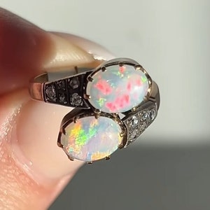 May include: A gold ring with two oval opals, one with a pink and white pattern and the other with a rainbow iridescent pattern. The ring is set with small diamonds around the opals.