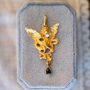 May include: A gold-coloured pendant featuring an angel design with outstretched wings and a small, clear gemstone. A black teardrop-shaped stone hangs from the bottom. The pendant is displayed on a grey velvet surface.