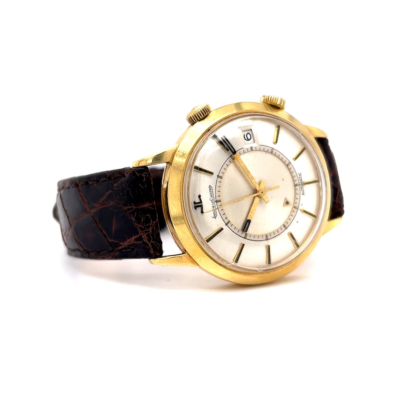 May include: A gold-tone wristwatch with a white face and black hands. The watch has a brown leather band and a date window at the 3 o'clock position. The watch face also has the text "Jaeger-LeCoultre Automatic" printed on it.
