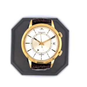 May include: A gold-tone wristwatch with a white face and black hands. The watch has a brown leather band and the words "Jaeger-LeCoultre" and "Automatic" printed on the face.