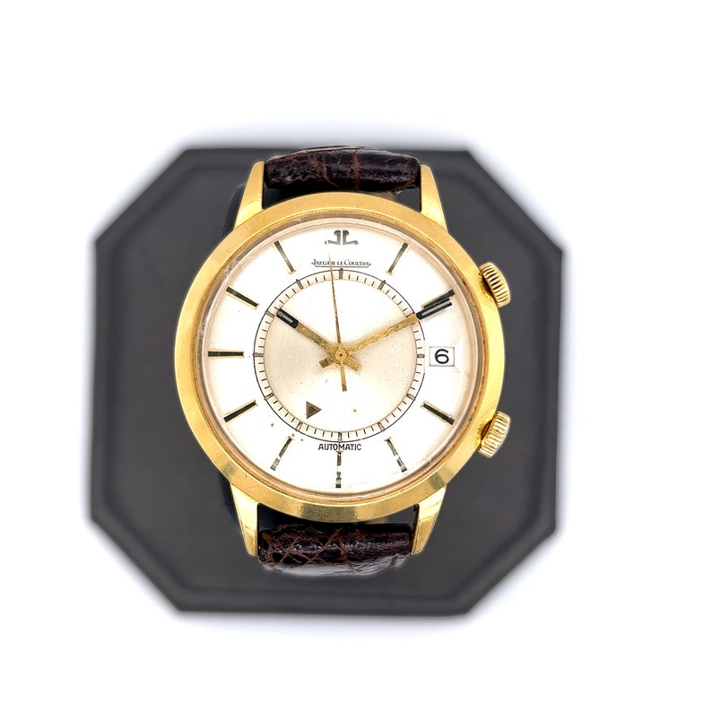 May include: A gold-tone wristwatch with a white face and black hands. The watch has a brown leather band and the words "Jaeger-LeCoultre" and "Automatic" printed on the face.