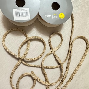 May include: Two spools of Spritz trim with a braided gold cord. The trim is labeled as 9 ft (2.7 m). The spools are gray with a white label and a black center hole. The cord is looped on a light beige surface.