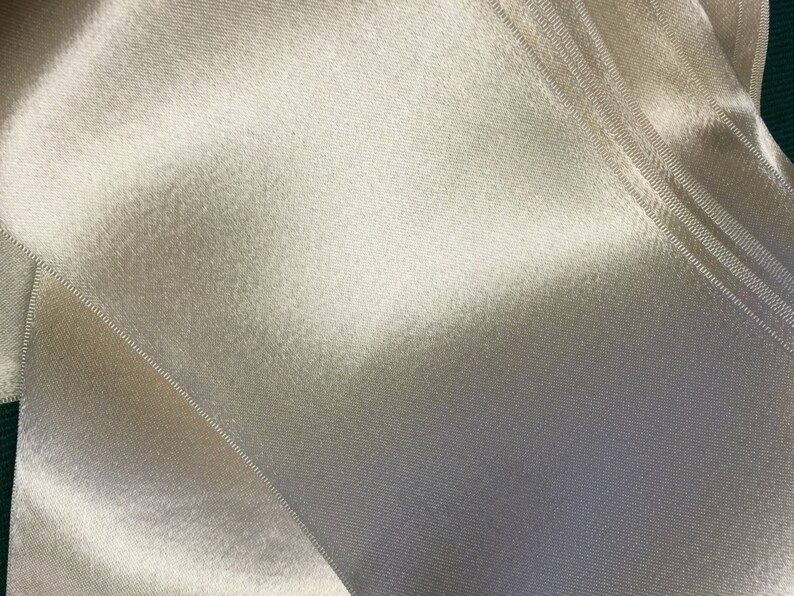 May include: A close-up of a white satin ribbon with a smooth, silky texture. The ribbon is folded in a way that creates a diagonal pattern.