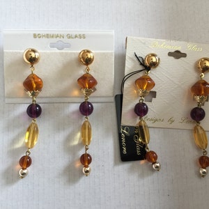 May include: A pair of gold dangle earrings with amber and purple glass beads. The earrings are labelled "Bohemian Glass Designs by Lenore".