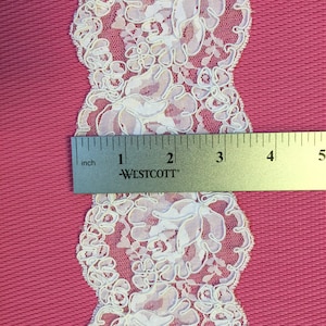 Op de afbeelding: Wit kantlint met een bloemenpatroon. De kant is ongeveer 1 inch breed. Een liniaal is opgenomen in de afbeelding voor schaal.