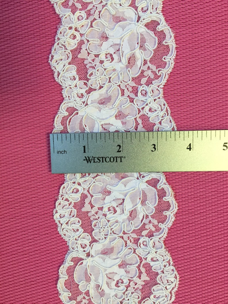 Op de afbeelding: Wit kantlint met een bloemenpatroon. De kant is ongeveer 1 inch breed. Een liniaal is opgenomen in de afbeelding voor schaal.