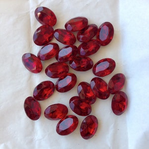 May include: A collection of oval-shaped, red, faceted glass gems. The gems are scattered on a white surface.