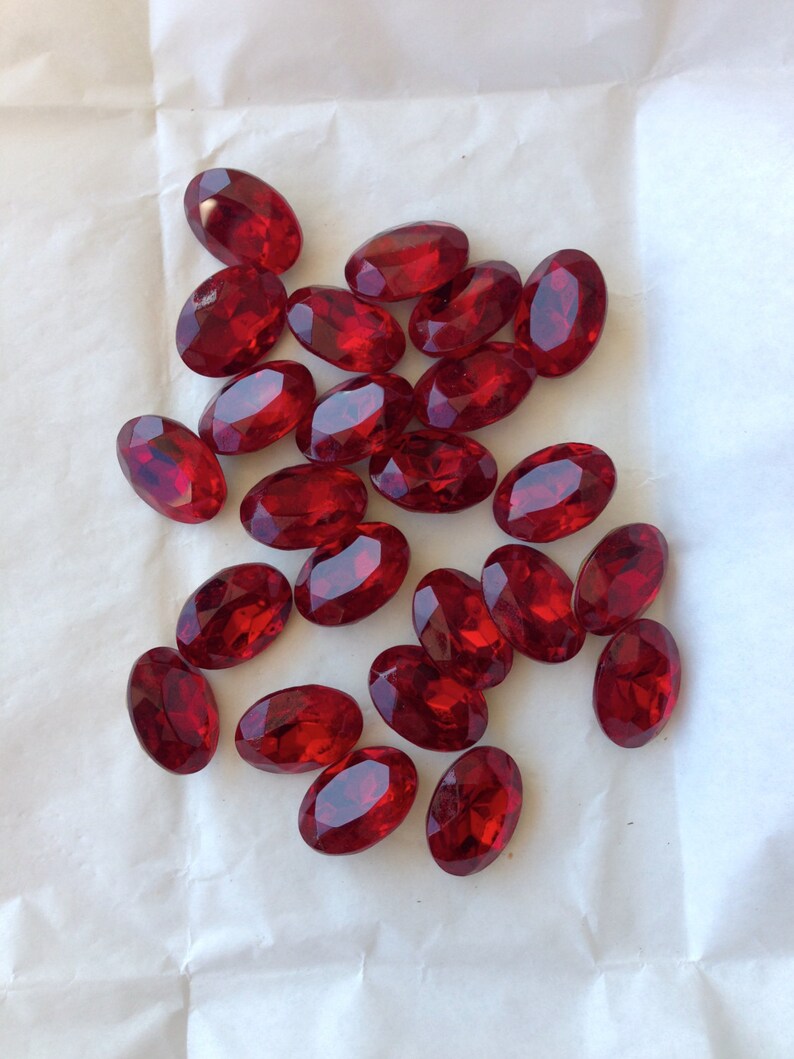 May include: A collection of oval-shaped, red, faceted glass gems. The gems are scattered on a white surface.