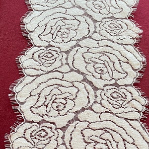May include: Cream lace table runner with a repeating rose design. The runner is laid on a burgundy surface. The edges of the runner have a scalloped trim.