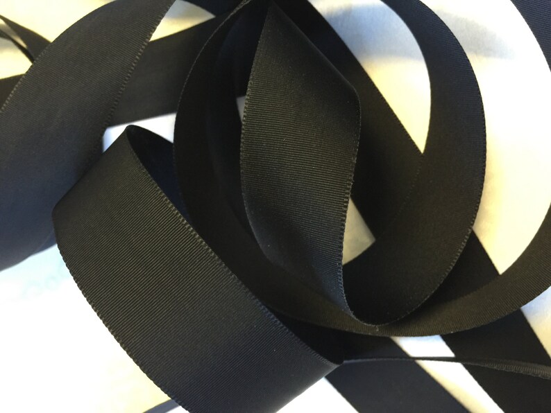 May include: Black grosgrain ribbon, a type of fabric ribbon with a textured, woven surface. The ribbon is folded and arranged in a decorative way.