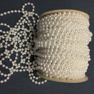 Puede incluir: Un carrete de ribete de perlas blancas, con un tramo del ribete desenrollado. El ribete está hecho de pequeñas perlas blancas redondas enhebradas en un alambre fino.
