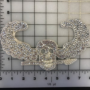 Puede incluir: Una calavera plateada con alas y un diseño de huesos cruzados, hecha de pedrería. La calavera mide aproximadamente 5 cm de ancho.
