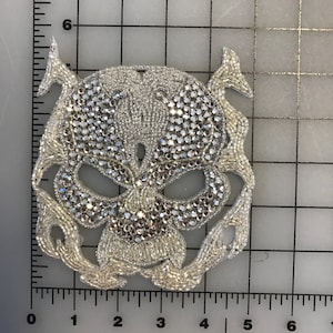Puede incluir: Un parche de calavera de plata con pedrería y abalorios. La calavera tiene un diseño detallado con una parte superior puntiaguda y una boca ancha y abierta.