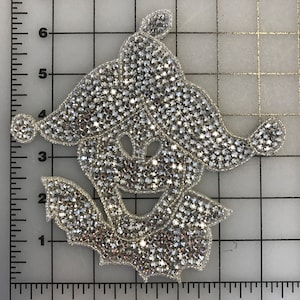 Puede incluir: Un parche de hierro de strass plateado en forma de bufón con una expresión caprichosa. El parche mide aproximadamente 10 cm de ancho y 7,5 cm de alto.