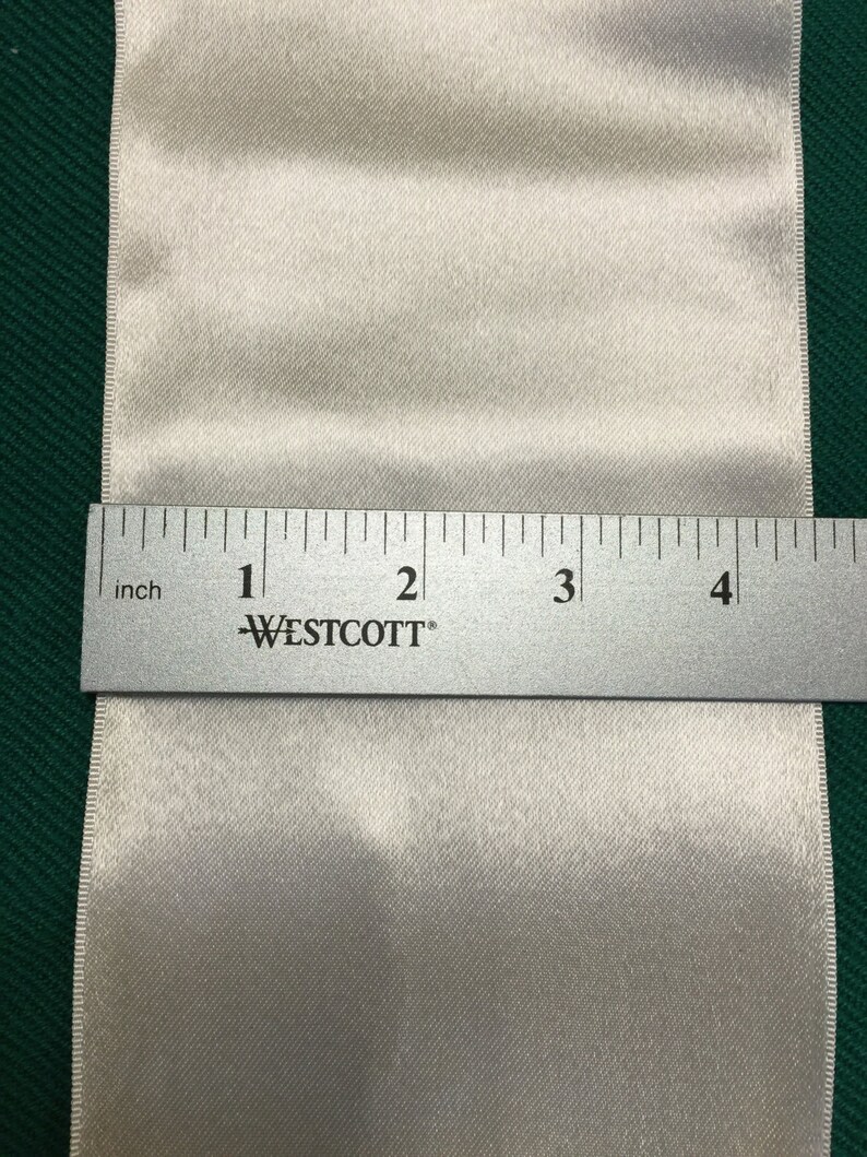 May include: A close-up of a white satin ribbon with a ruler measuring 4 inches in length. The ruler is labeled "WESTCOTT" and has markings for inches.