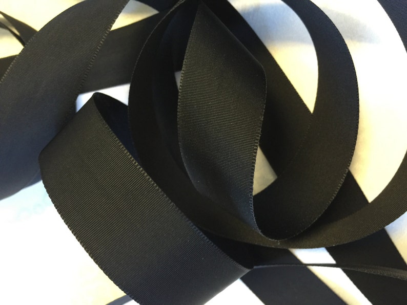 May include: Black grosgrain ribbon, a type of fabric ribbon with a textured, woven surface. The ribbon is folded and arranged in a decorative way.