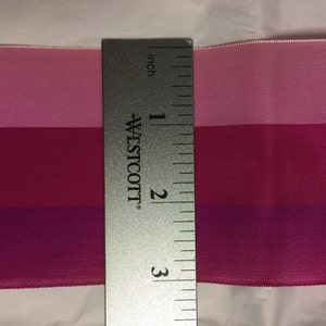 Puede incluir: Una cinta de tres tonos rosa, rojo y morado medida con una regla. La cinta mide 7,6 cm de largo.