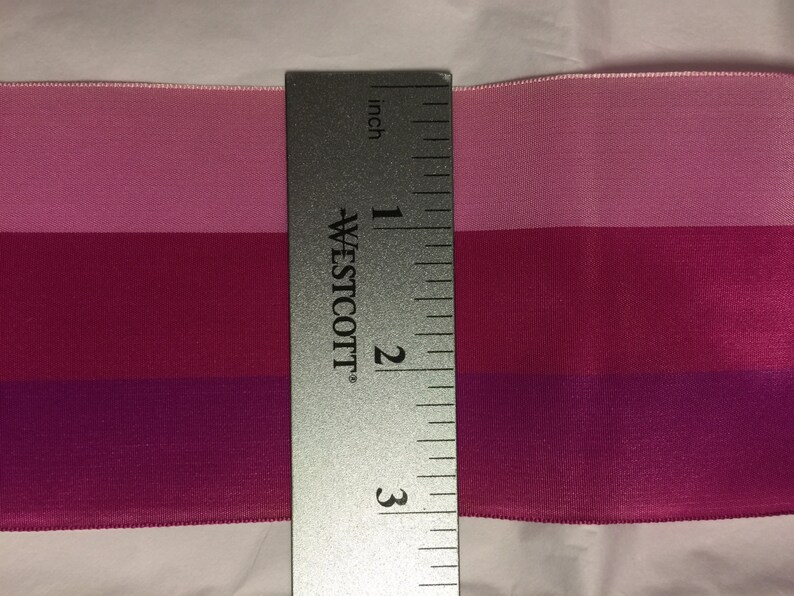 Puede incluir: Una cinta de tres tonos rosa, rojo y morado medida con una regla. La cinta mide 7,6 cm de largo.