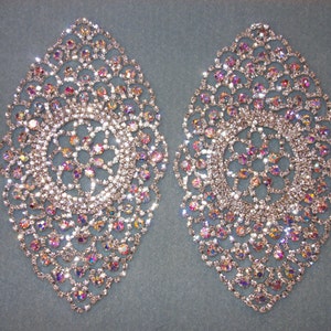 May include: Two silver-toned rhinestone embellishments in a teardrop shape with a circular design in the center. The rhinestones are iridescent and sparkle.