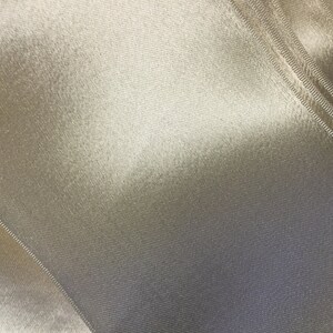 May include: Close-up of a smooth, off-white fabric with a subtle sheen. The fabric has a fine, woven texture and a slight crinkle.