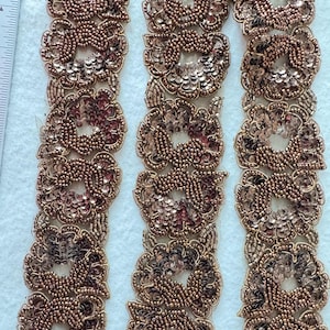 Puede incluir: Tres tiras de ribete de cuentas doradas con lentejuelas. El ribete presenta un patrón repetitivo de formas florales.