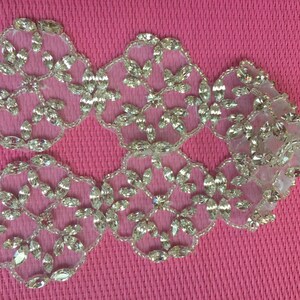 May include: Close-up of several clear, sparkling rhinestone appliques arranged on a pink textured surface. Each applique features a floral design with multiple clear rhinestones and a beaded border. The appliques are likely for embellishing garments or accessories.
