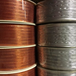 K&ouml;nnte beinhalten: Rollen von orangefarbenem und silbernem Band mit schimmerndem Finish. Das Band ist um Spulen gewickelt und &uuml;bereinander gestapelt.