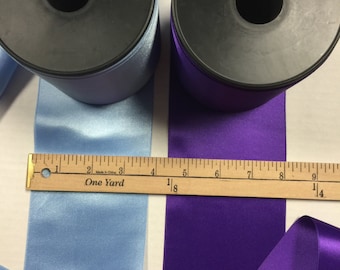 May include: Two spools of satin ribbon, one light blue and one purple. The spools are labelled with the brand name "SENSUEL" and the text "0422 SATIN T.TEXTILES 095 mm x 25 M cal 231 100% POLYESTER". A ruler is placed below the spools, showing the length of the ribbon.