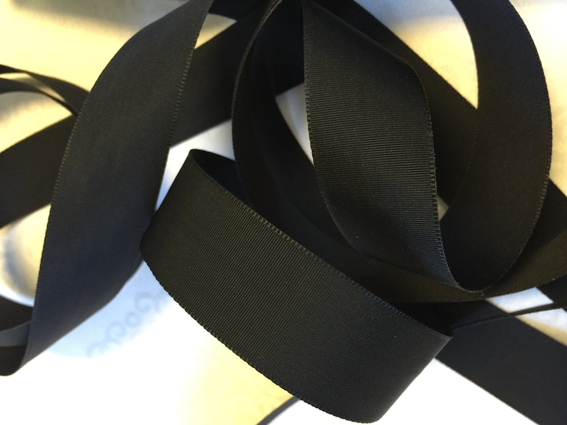 May include: Black grosgrain ribbon, a type of fabric ribbon with a textured, woven surface. The ribbon is folded and twisted, showing its width and texture.