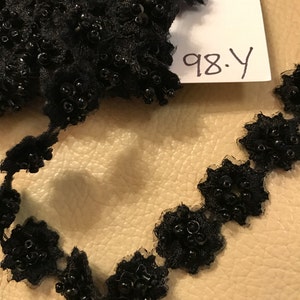 以下が含まれることがあります： 黒いビーズレーストリム、花柄のデザイン。トリムは小さくて丸い黒いビーズと繊細なスカラップエッジが特徴です。「98.Y」と手書きされた白いタグが見えます。