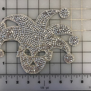 Puede incluir: Una aplicación de sombrero y máscara de bufón de pedrería plateada. El diseño está hecho de pequeñas piedras de pedrería redondas.