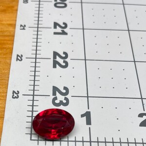 May include: A deep red oval-shaped gemstone measuring approximately 1 inch in length. The gemstone is sitting on a white gridded ruler.